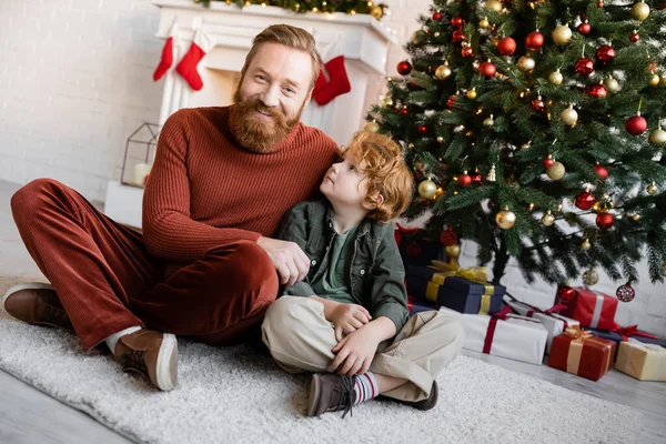 Pelirroja mirando sonriente papá barbudo en el suelo en la sala de estar cerca de cajas de regalo bajo el árbol de Navidad - foto de stock