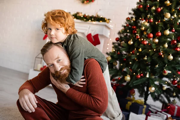 Happy bearded man looking at camera while piggybacking redhead son near Christmas tree — Stock Photo
