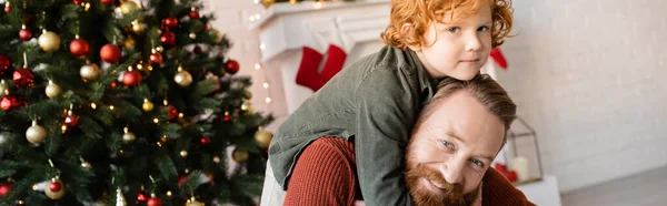 Feliz barbudo hombre piggybacking pelirroja hijo cerca borrosa árbol de Navidad, pancarta - foto de stock