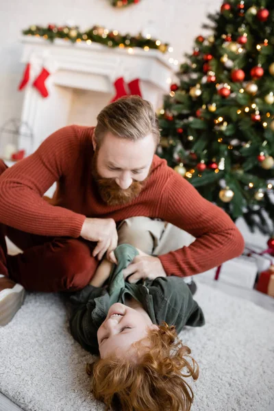 Redhead boy laughing on floor while having fun with dad near blurred Christmas tree — Stock Photo