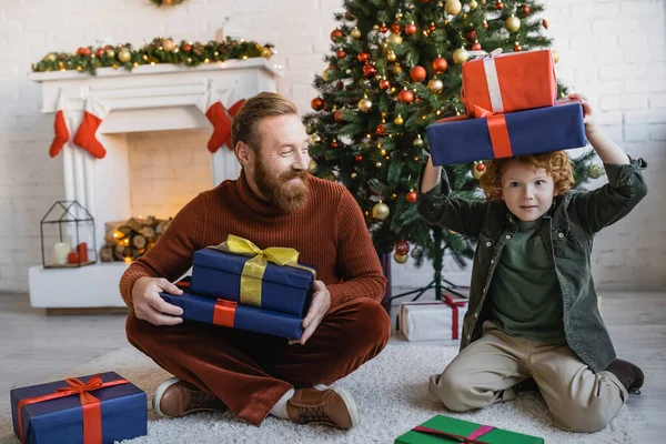 Niño sosteniendo cajas de regalo por encima de la cabeza mientras se divierte cerca de papá barbudo sentado en el suelo cerca del árbol de Navidad - foto de stock