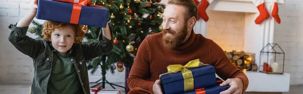 Улыбающийся бородатый мужчина смотрит на рыжего сына с подарочной коробкой над головой во время празднования Рождества, баннер — стоковое фото