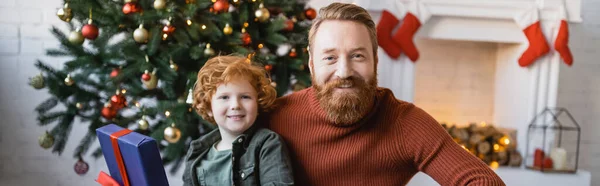 Alegre e ruiva pai e filho olhando para a câmera na sala de estar com árvore de Natal e lareira, banner — Fotografia de Stock