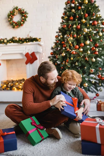 Alegre ruiva menino segurando presente de Natal enquanto sentado no chão perto de pai barbudo na sala de estar decorada — Fotografia de Stock