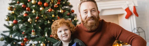 Felice rossa ragazzo con il padre barbuto guardando la fotocamera vicino all'albero di Natale decorato, banner — Foto stock