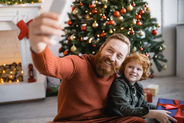 Cheerful bearded man taking selfie with redhead son holding gift box near Christmas tree — Stock Photo