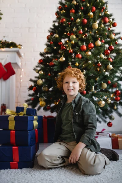 Menino ruivo sentado no chão perto de presentes de Natal e pinheiros decorados com bugigangas em casa — Fotografia de Stock