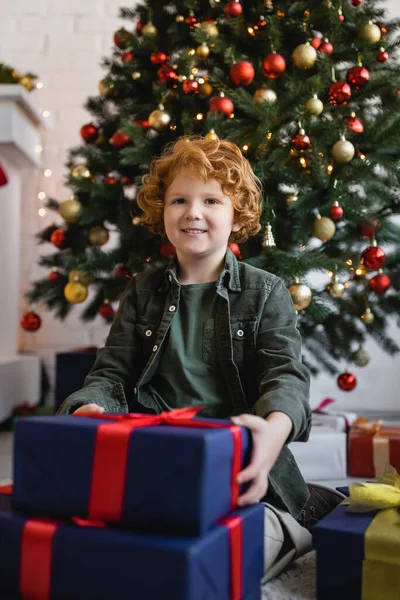 Niño alegre con el pelo rojo sonriendo a la cámara cerca de regalos de Navidad y pino decorado - foto de stock
