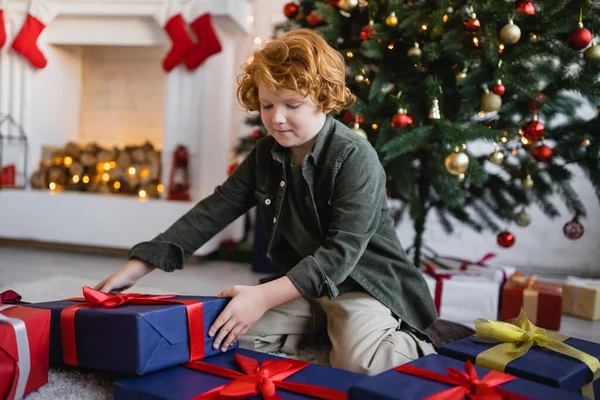 Criança ruiva sentada no chão perto de presentes de Natal na sala de estar decorada no fundo borrado — Fotografia de Stock