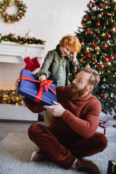 Pelirroja dando regalo de Navidad a papá sorprendido sentado en el suelo con las piernas cruzadas - foto de stock