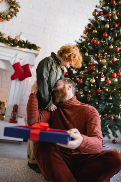 Feliz hombre sentado en el suelo con regalo de Navidad y abrazando pelirroja hijo - foto de stock