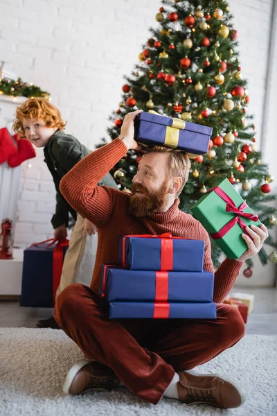Веселый бородатый мужчина веселится с подарочными коробками рядом с сыном и елкой на размытом фоне — стоковое фото