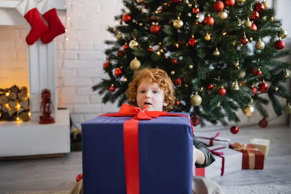 Asombrado pelirrojo chico mirando la cámara detrás de enorme caja de regalo y árbol de Navidad en sala de estar - foto de stock