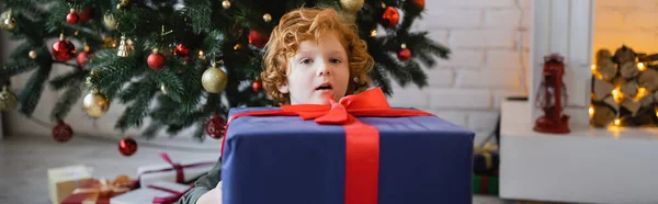 Bambino rosso stupito guardando la fotocamera vicino alla grande scatola regalo e all'albero di Natale, banner — Foto stock