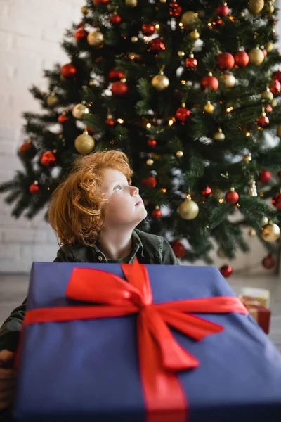 Criança ruiva olhando para longe perto enorme caixa de presente e árvore de Natal decorada — Fotografia de Stock