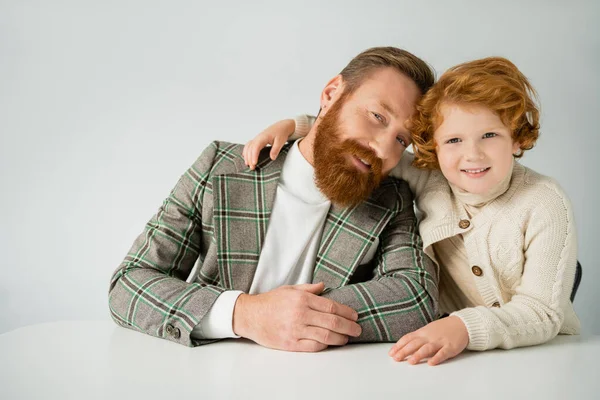 Redhead boy hugging dad in plaid blazer and smiling at camera while lying on grey background — Stock Photo