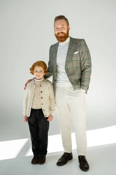 Volle Länge des trendigen bärtigen Mannes, der mit der Hand in der Tasche neben dem rothaarigen Sohn auf grauem Hintergrund steht — Stockfoto