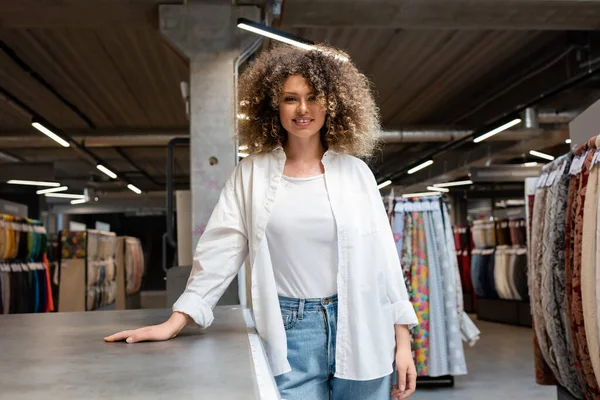 Cheerful and curly saleswoman standing near desk in textile shop — Stock Photo