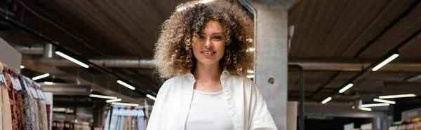 Cheerful and curly saleswoman smiling in textile shop, banner — Stock Photo