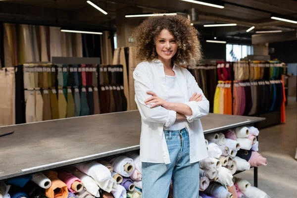 Cheerful and curly saleswoman standing with crossed arms near rack with different fabric rolls in textile shop — Stock Photo