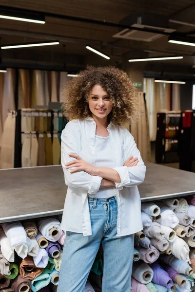 Glückliche Verkäuferin, die mit verschränkten Armen neben dem Regal mit verschiedenen Stoffrollen im Textilgeschäft steht — Stockfoto