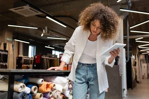 Curly saleswoman holding digital tablet and checking fabric rolls in textile shop — Stock Photo