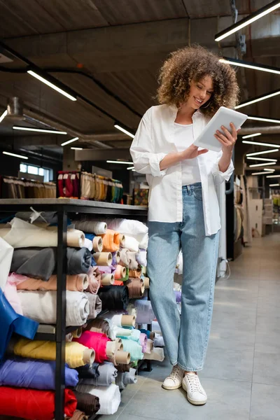 Zufriedene Verkäuferin mit digitalem Tablet in der Nähe von Regalen mit Stoffrollen im Textilgeschäft — Stockfoto