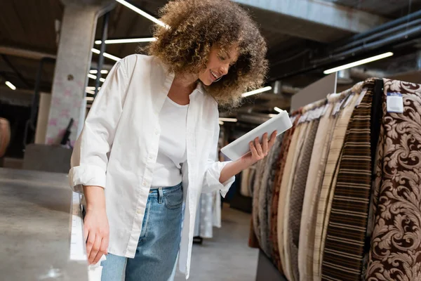 Zufriedene Verkäuferin mit digitalem Tablet in der Nähe von gemustertem Stoff im Textilgeschäft — Stockfoto