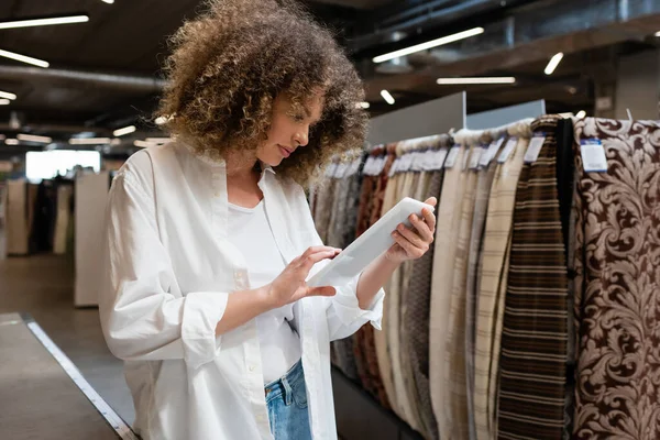 Fokussierte und lockige Verkäuferin mit digitalem Tablet in der Nähe gemusterter Stoffe im Textilgeschäft — Stockfoto