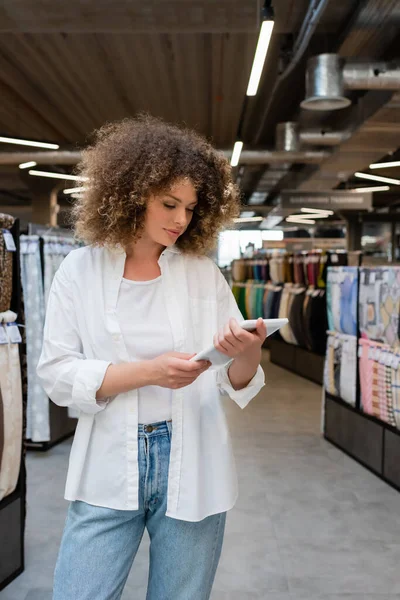 Vendedora encaracolado usando tablet digital perto de tecido diferente na loja têxtil — Fotografia de Stock