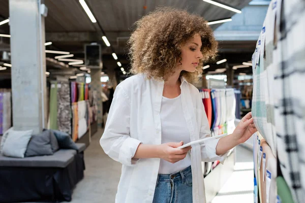 Seitenansicht der lockigen Verkäuferin, die ein digitales Tablet in der Hand hält, während sie im Textilgeschäft karierte Stoffe prüft — Stockfoto