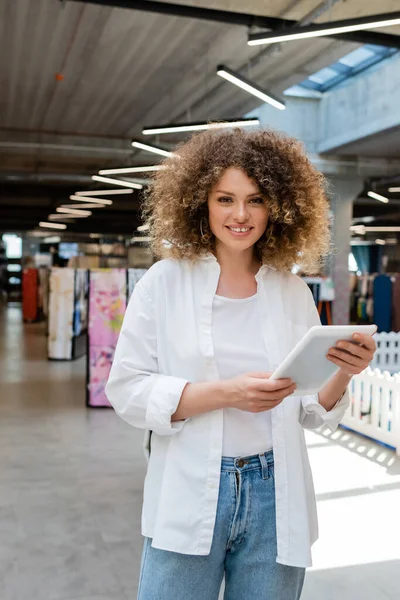 Vendeuse souriante aux cheveux bouclés tenant une tablette numérique dans un magasin de textiles — Photo de stock