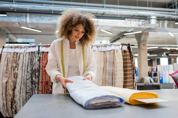 Smiling saleswoman with measuring tape holding blue linen fabric roll in textile shop — Stock Photo
