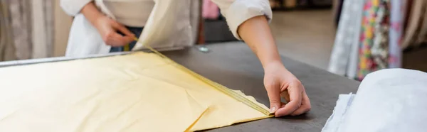 Обрезанный вид продавщицы измеряющей желтую льняную ткань в текстильном магазине, баннер — стоковое фото
