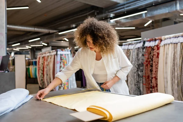 Vendedor joven y rizado que mide el rollo de tela de lino amarillo en la tienda textil - foto de stock