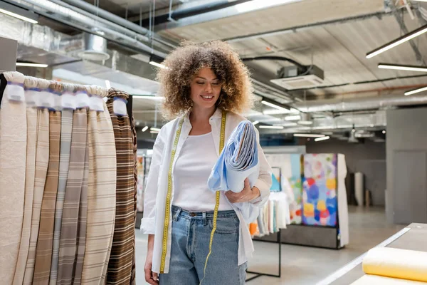 Heureuse vendeuse avec ruban à mesurer portant rouleau de tissu bleu dans la boutique textile — Photo de stock