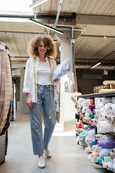 Fröhliche Verkäuferin mit Maßband trägt blaue Stoffrolle in Textilladen — Stockfoto
