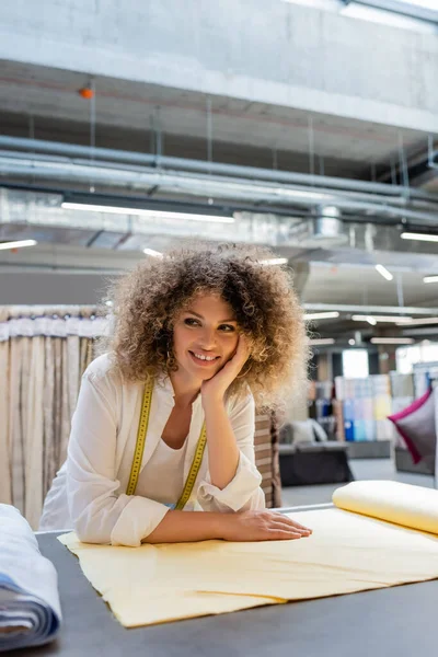 Heureuse vendeuse avec ruban à mesurer souriant près des rouleaux de tissu dans la boutique textile — Photo de stock