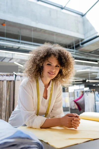 Joyful saleswoman with measuring tape smiling near fabric rolls in textile shop — Stock Photo