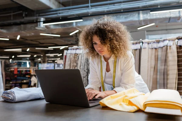 Curly saleswoman with measuring tape using laptop near fabric rolls in textile shop — Stock Photo