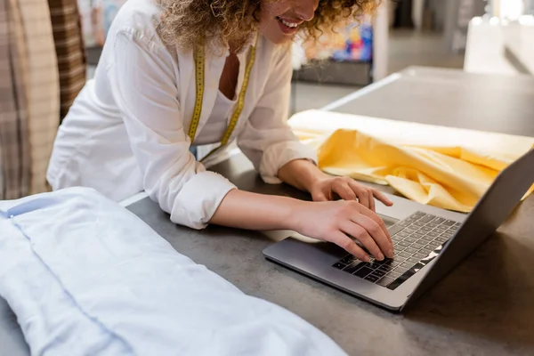 Abgeschnittene Ansicht der lockigen Verkäuferin lächelnd und mit Laptop in der Nähe von Stoffrollen im Textilgeschäft — Stockfoto