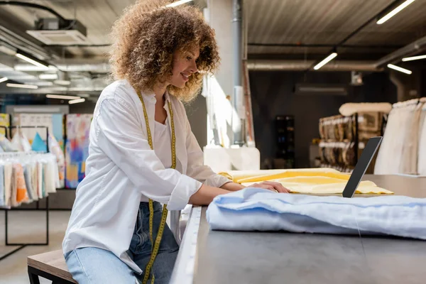 Side view of happy saleswoman with measuring tape using laptop near fabric rolls in textile shop — Stock Photo