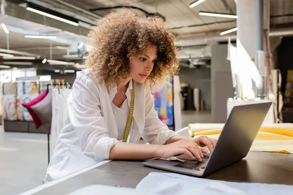 Curly and young saleswoman using laptop while working in textile shop — Stock Photo