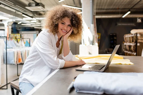 Vendedora encaracolado com fita métrica sorrindo perto de laptop e rolos de tecido na loja têxtil — Fotografia de Stock