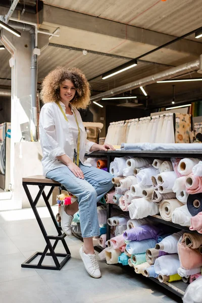Happy saleswoman with curly hair sitting on high chair near rack with fabric rolls in textile shop — Stock Photo