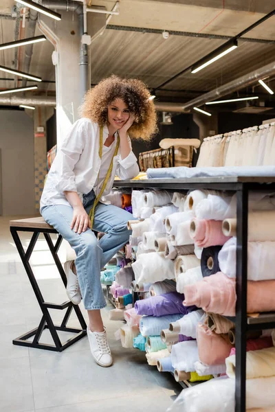 Fröhliche Verkäuferin mit Maßband sitzt auf Hochstuhl in der Nähe von Gestell mit Stoffrollen im Textilgeschäft — Stockfoto
