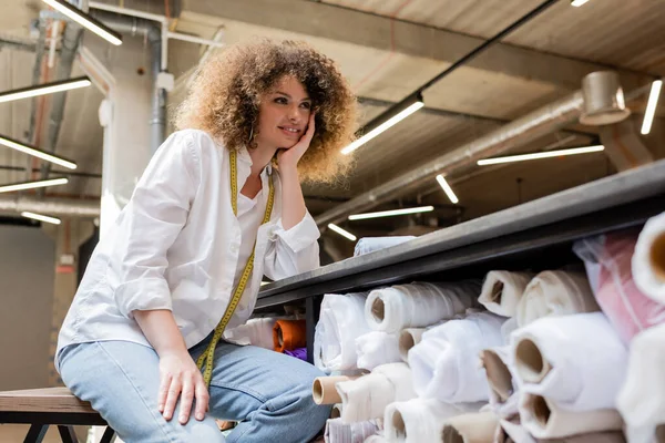 Cheerful saleswoman with measuring tape sitting on chair near rack with fabric rolls in textile shop — Stock Photo