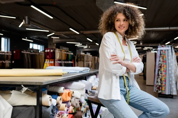 Joyful saleswoman with curly hair posing with crossed arms near rack with fabric rolls in textile shop — Stock Photo