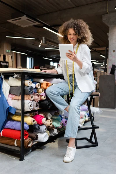 Jubilosa vendedora sentada em cadeira alta e usando tablet digital perto de rack com rolos de tecido na loja têxtil — Fotografia de Stock