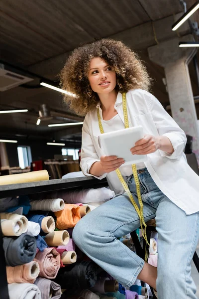Happy saleswoman sitting on chair and holding digital tablet near rack with fabric rolls in textile shop — Stock Photo